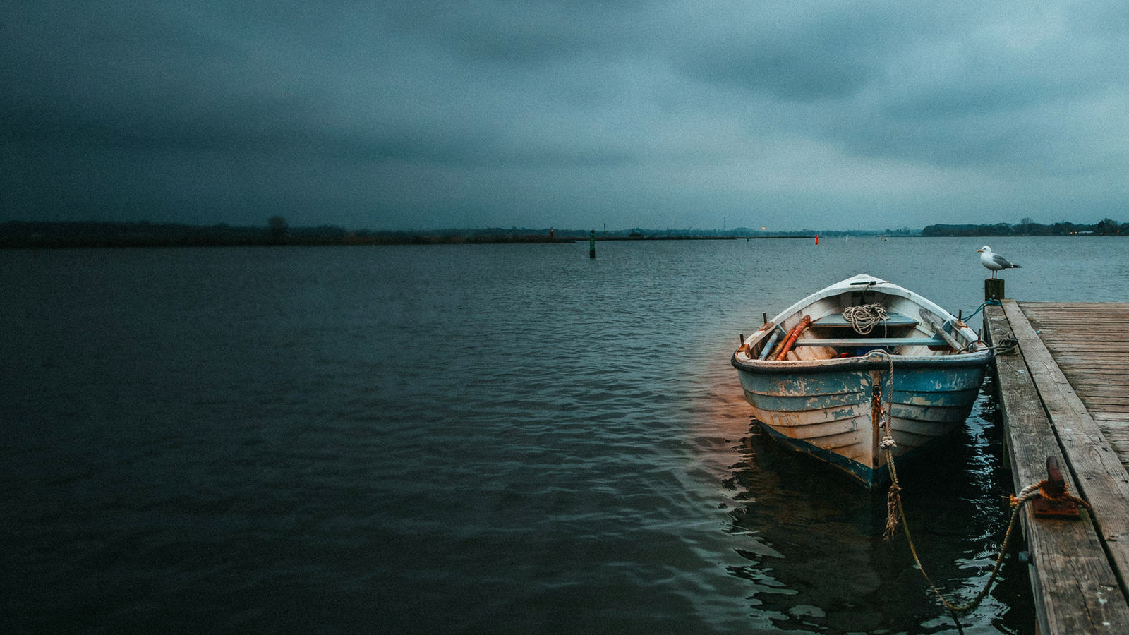 A weathered wooden boat tied to a dock in a still harbor at dusk under a brooding grey sky, the ship kept in the harbor just in case
