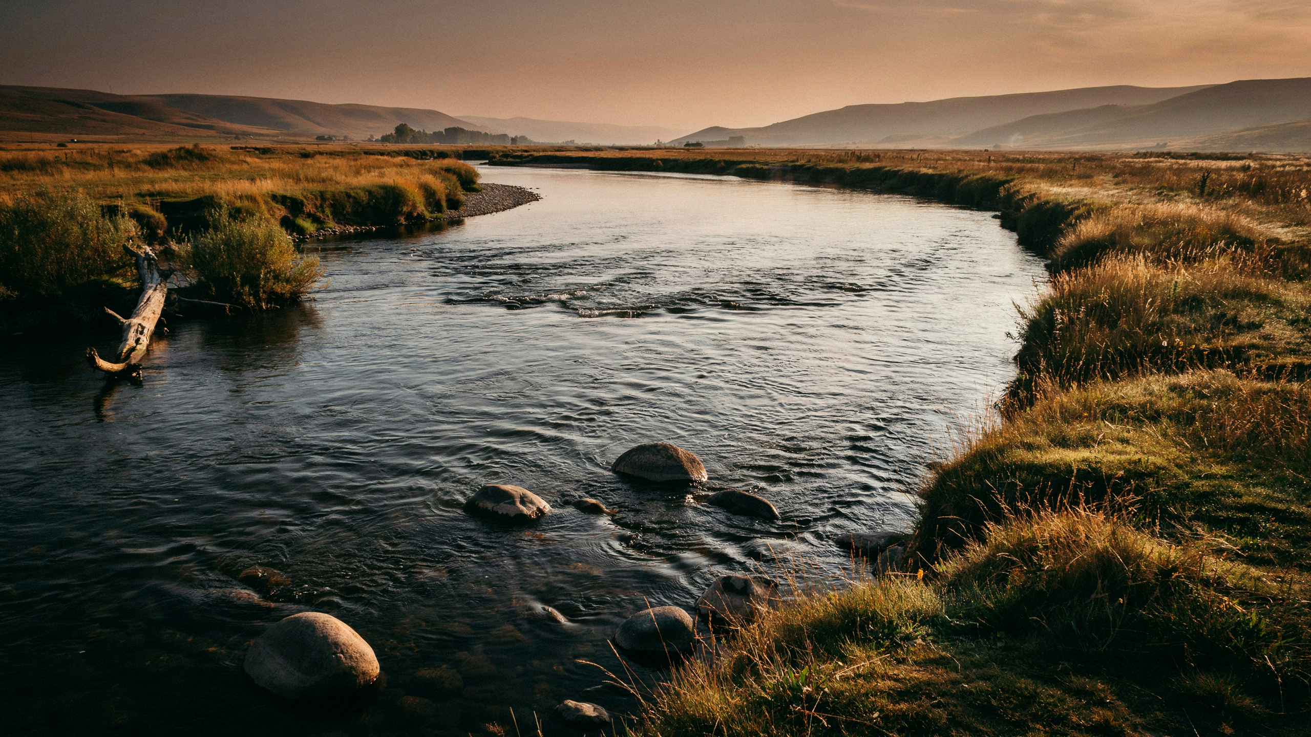 A wide river bending gently through open landscape at dawn, warm copper light on moving water with visible ripples over stones in the current