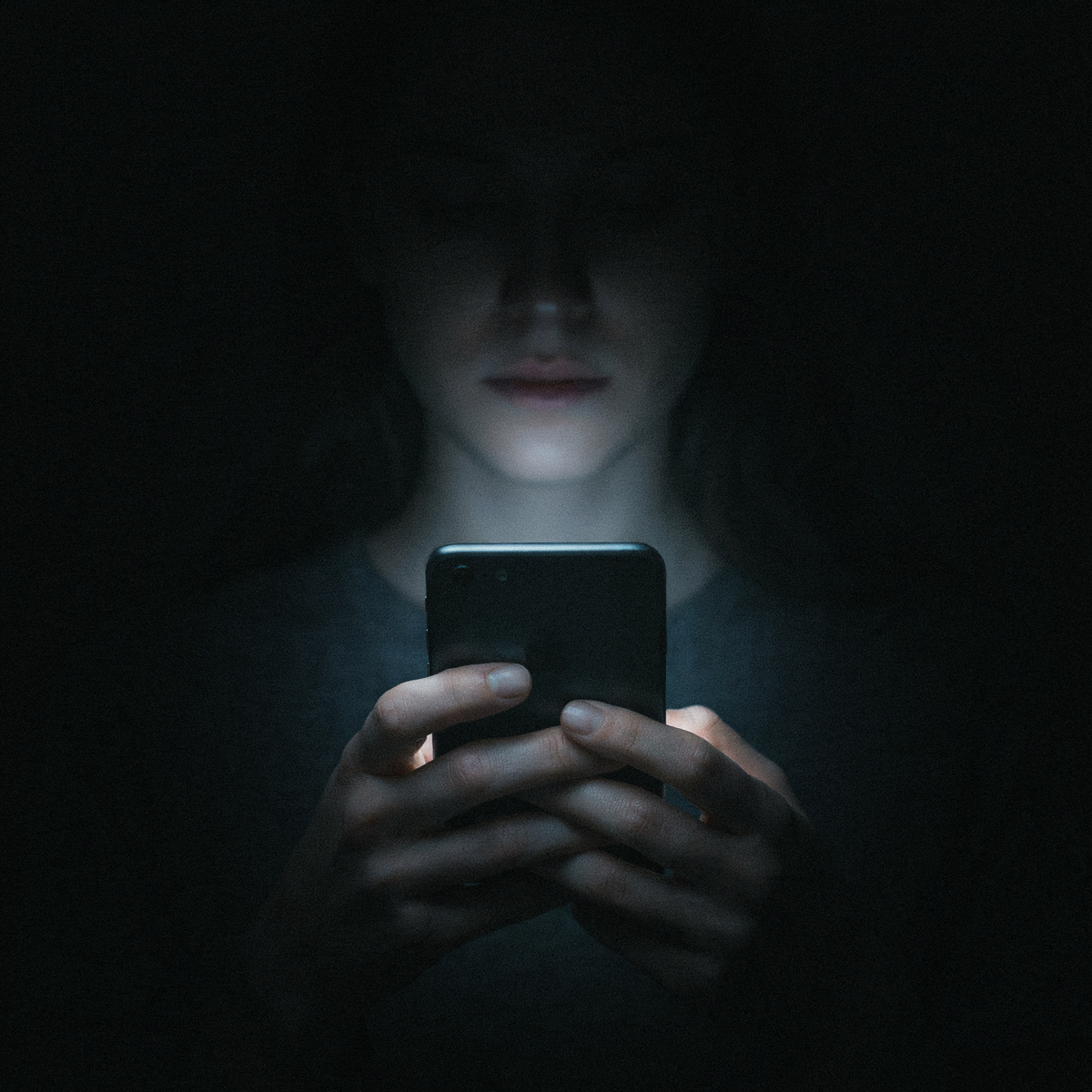 A single hand holding a smartphone at night, the screen's cold glow underlighting the hand in deep black surroundings