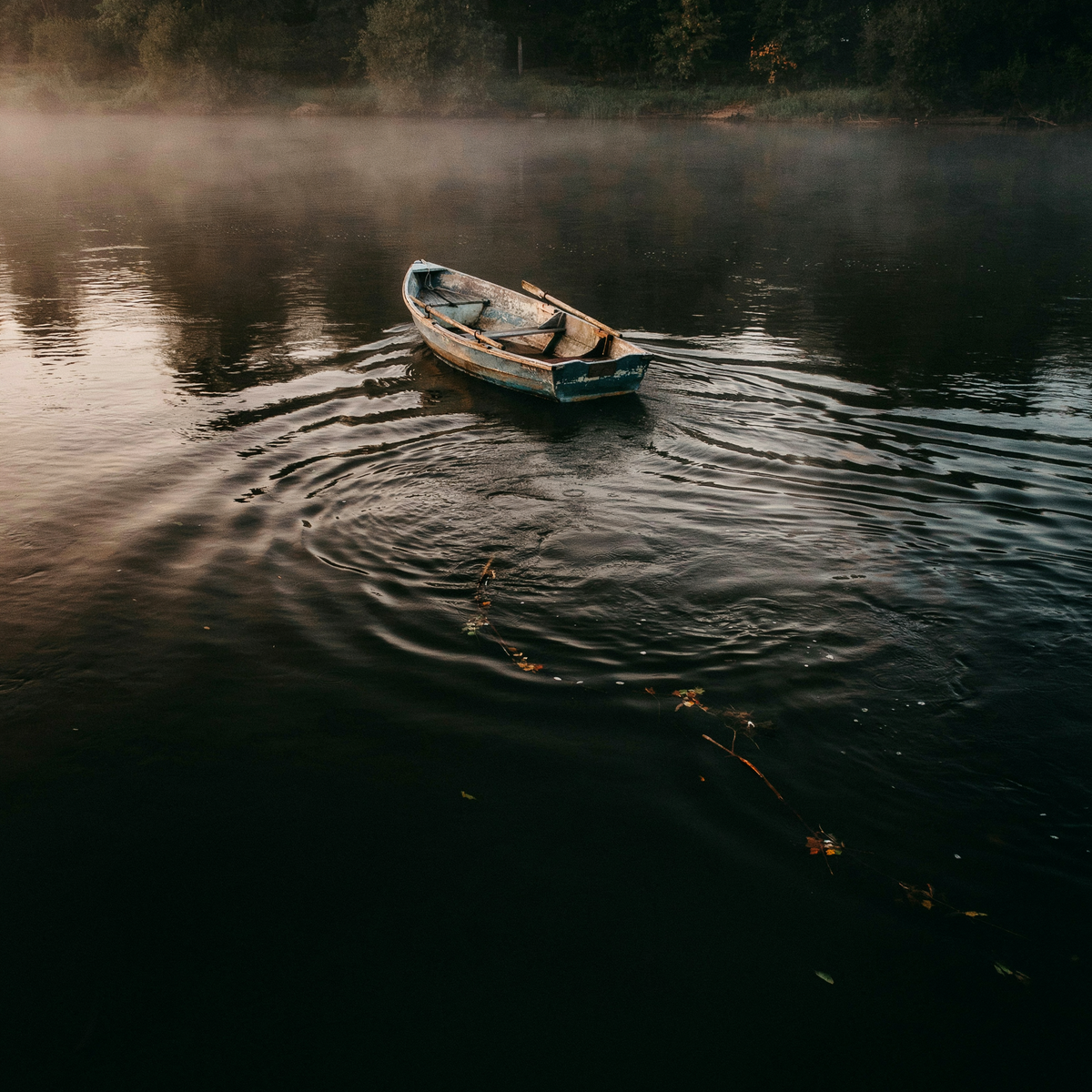 An empty wooden rowboat caught sideways in dark river currents at dusk, fog rolling over the water with foggy trees in the distance, copper-orange light catching the rippled surface as concentric waves radiate from the vessel pulled off course