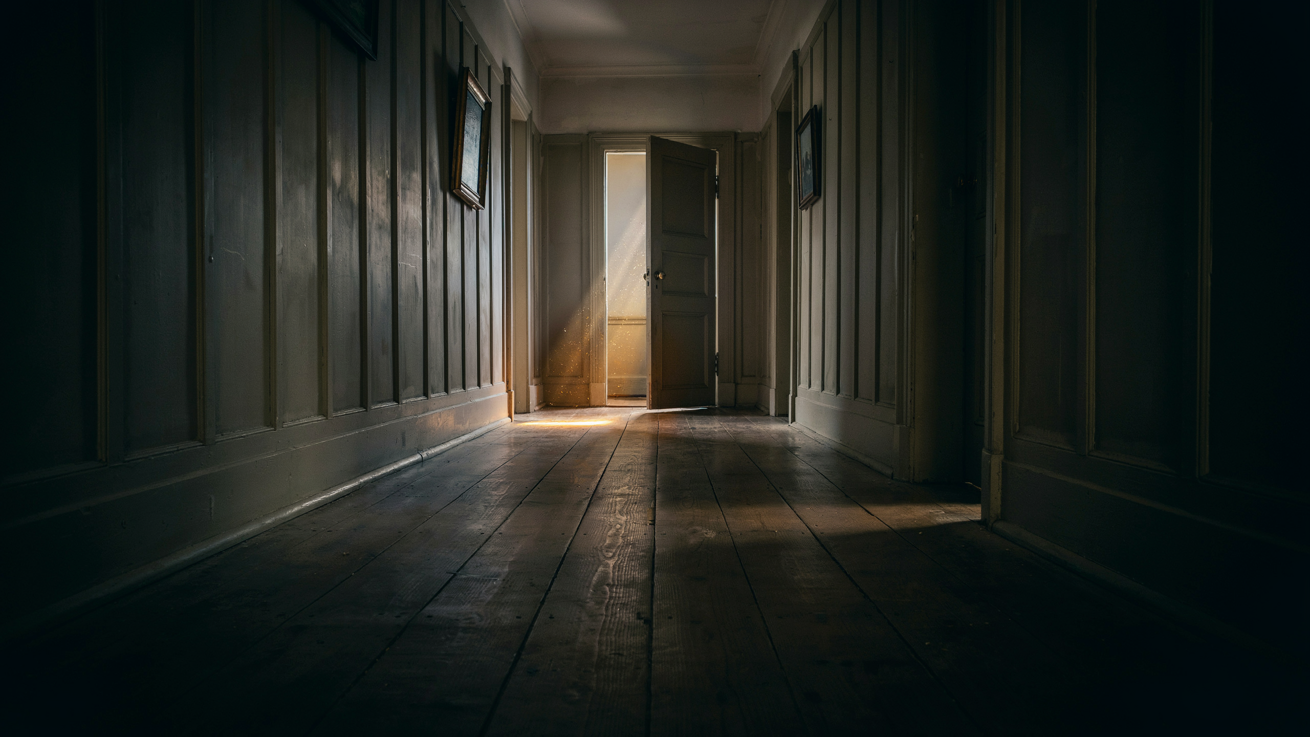 A long dark hallway with a single partially open door at the far end pouring warm golden light across the worn floorboards, the door that only opens from the inside