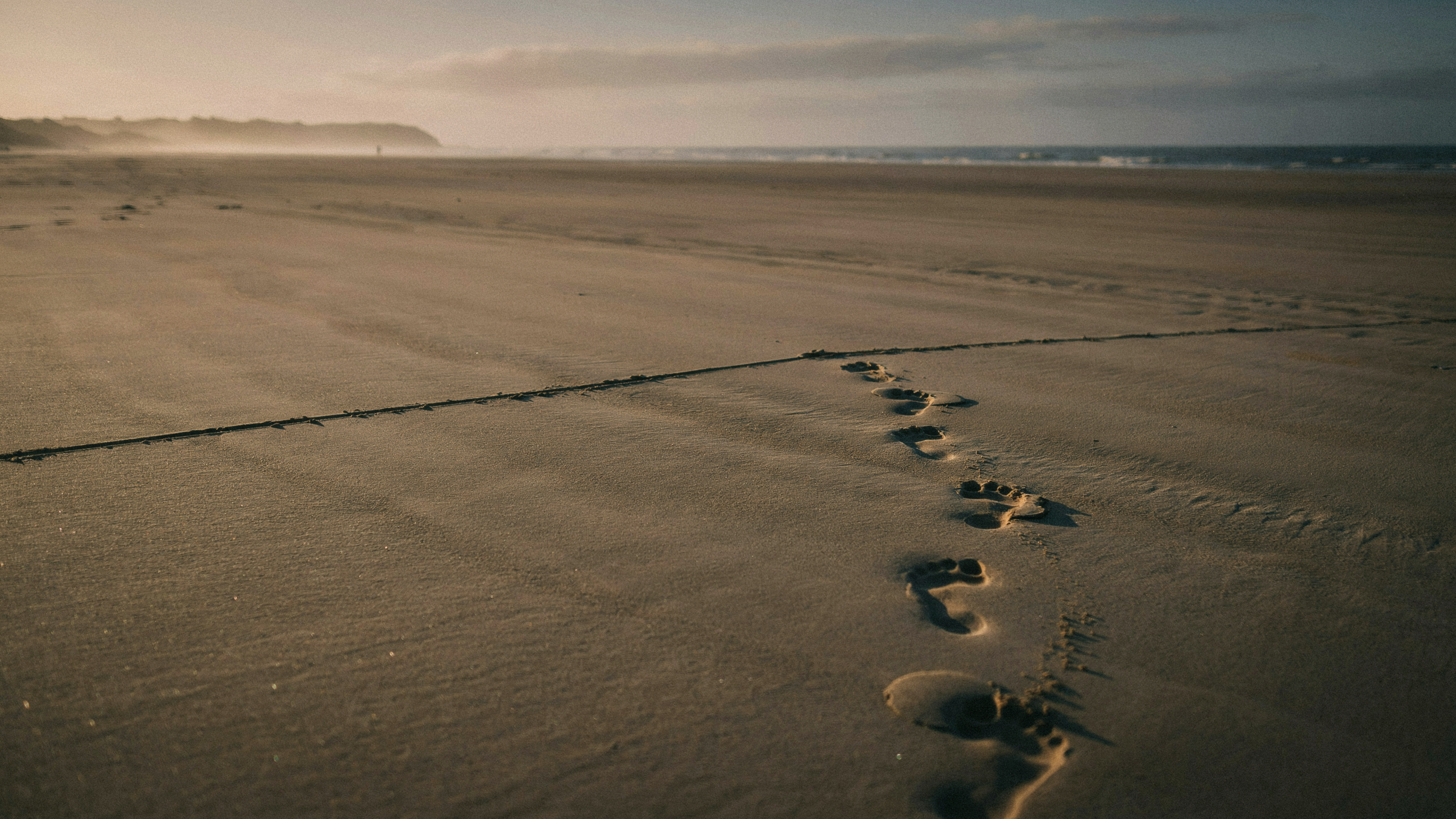 A single line drawn across a wide stretch of soft sand at golden hour, footprints leading up to it but not crossing, the moment of pause at the threshold